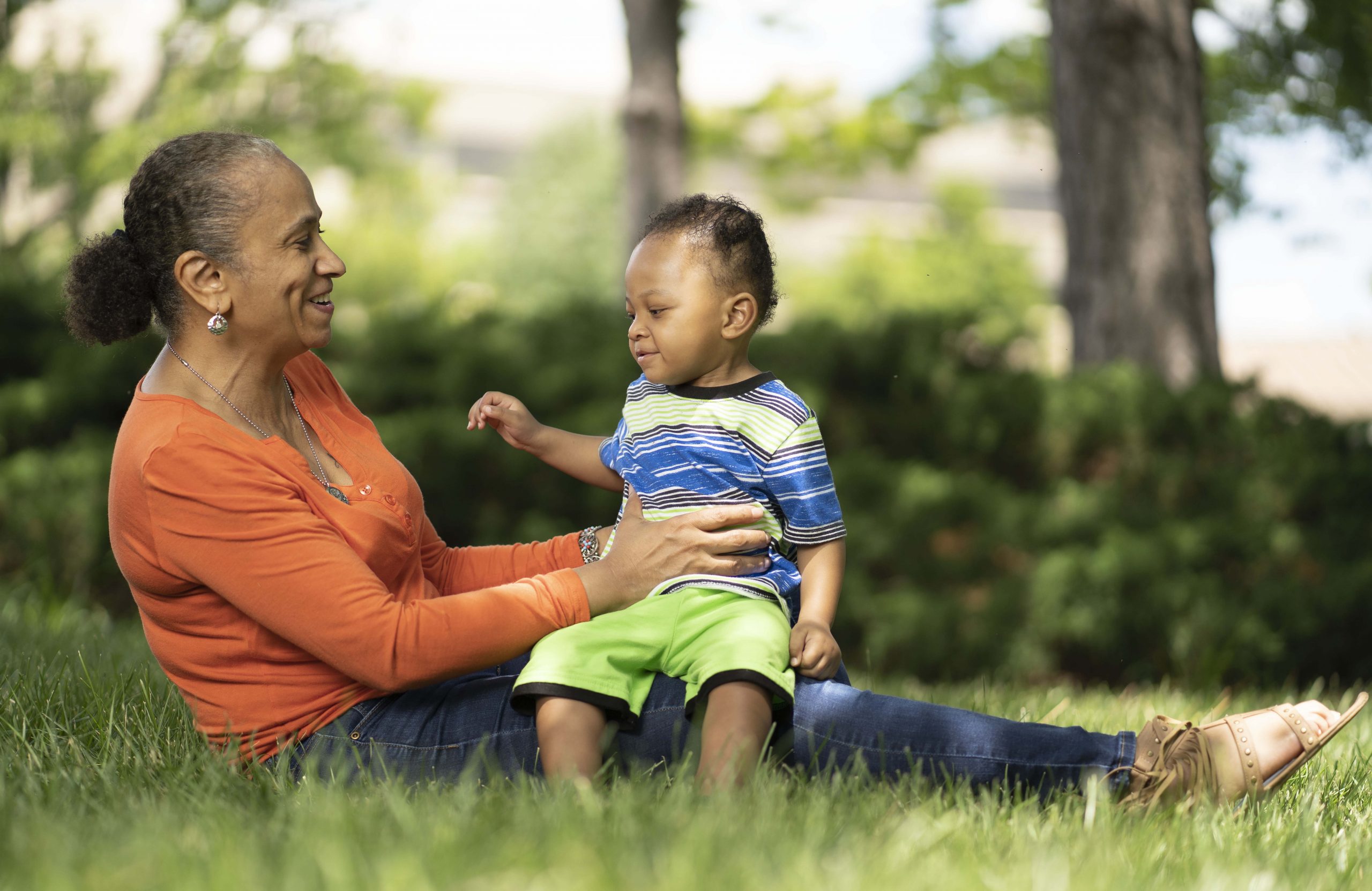 Britepaths' Client Eva sitting in the grass with her toddler grandson in her lap. They are both smiling.